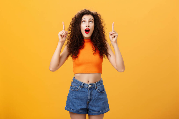 Portrait of amazed emotive and charismatic young curly-haired woman with red lipstic looking and pointing up mesmerized and astonished standing under impression over orange background - Photo, Image