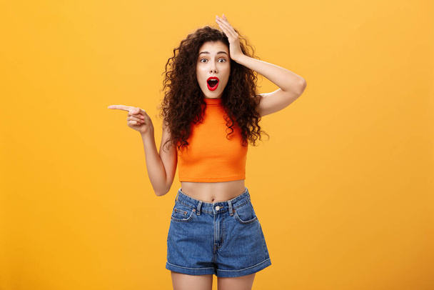 Surprised thrilled beautiful caucasian girl with curly hairstyle in red lipstic and orange cropped top holding hand on head amazed and pointing left questioned and astonished over studio background - Photo, Image