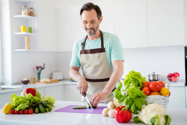 Photo of shiny attractive mature guy wear blue t-shirt apron smiling cooking cutting vegetables indoors house room - Valokuva, kuva