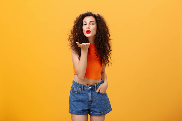 Stylish flirty and confident female with curly hairstyle and red lipstick smiling sensually bending towards camera sending gentle loving kiss at camera with palm near mouth posing near orange wall - Foto, immagini