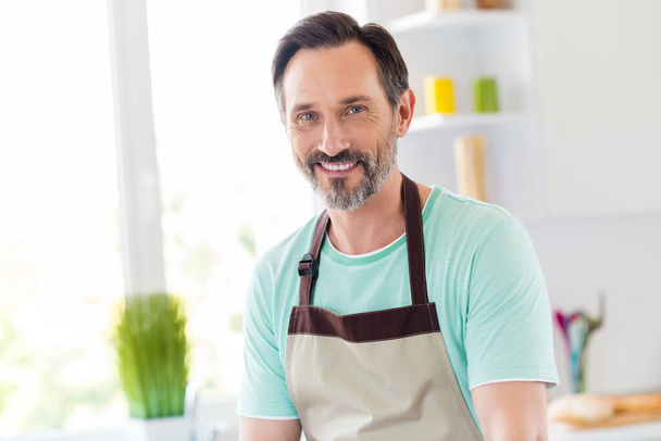 Zdjęcie przystojnego brodatego dojrzałego mężczyzny błyszczący uśmiech uśmiech nosić fartuch niebieski t-shirt domu kuchnia wewnątrz - Zdjęcie, obraz