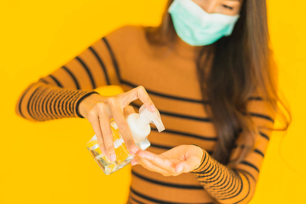 Retrato hermosa mujer asiática joven con la máscara de alcohol en aerosol y botella de gel en la mano para proteger contra el coronavirus o covid19 sobre fondo aislado amarillo
 - Foto, imagen