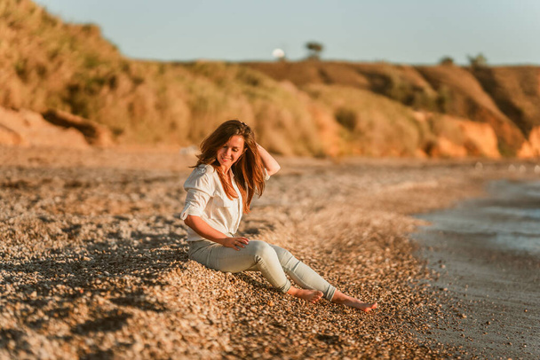  Чарівна молода жінка в білій сорочці і джинсах на спокійному романтичному пляжі біля моря. Концепція літніх жінок у відпустці. Поняття активного життя
. - Фото, зображення