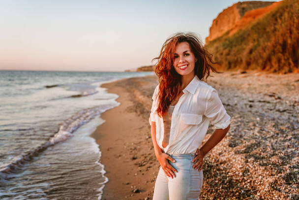  Μια γοητευτική νεαρή γυναίκα με λευκό πουκάμισο και τζιν σε μια ήρεμη ρομαντική παραλία δίπλα στη θάλασσα. Η έννοια των καλοκαιρινών διακοπών. Η έννοια της ενεργού ζωής. - Φωτογραφία, εικόνα