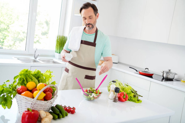 Photo of focused calm mature man cook salad add salt spices wear apron blue t-shirt home kitchen indoors - Foto, imagen