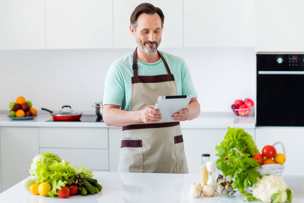 Fénykép a kíváncsi okos szakállas férfi hold tabletta olvasni recept kopás konyhai kötény a konyhában lakás beltéri - Fotó, kép