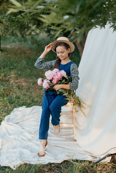 Outdoor portrait of young beautiful girl wearing blue suit, straw hat, holding peonies, walking in park. Spring, summer fashion concept - Fotó, kép