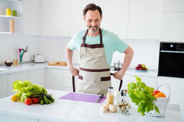 Photo of cheerful positive man prepare vegetarian meal toothy smile wear apron blue t-shirt home kitchen indoors - Valokuva, kuva