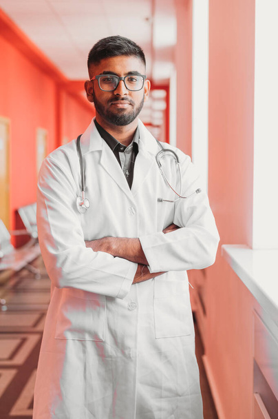 Hymyilevä intialainen lääkäri käyttää silmälaseja ja stetoskooppia valkoisessa takissa. Edessä näkymä muotokuva sairaalassa - Valokuva, kuva