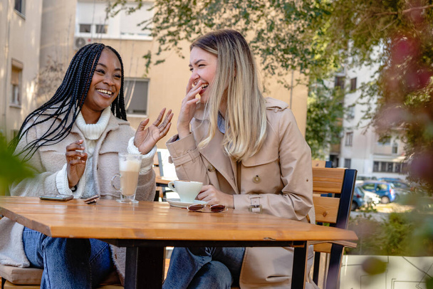 Twee jonge vriendinnen genieten samen van koffie op een terras van de coffeeshop, zitten aan een tafel, kletsen en lachen hardop. Diversiteit en multi-etnisch concept. - Foto, afbeelding