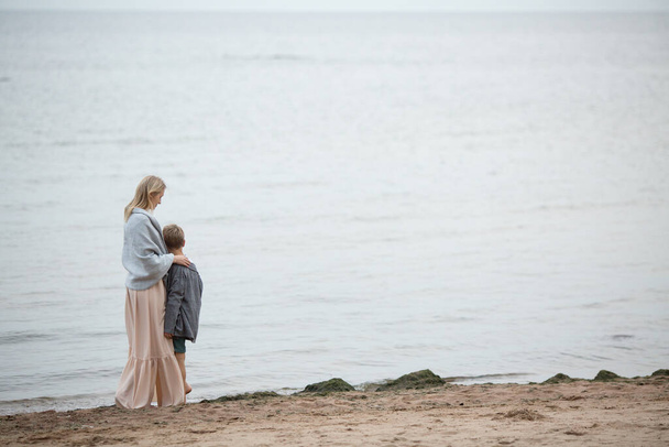 Une mère blonde dans une longue robe rose est enveloppée dans une couverture chaude, embrassant son jeune fils et le regardant, regardant la mer. Vue de l'arrière. Promenade estivale le long du rivage. - Photo, image
