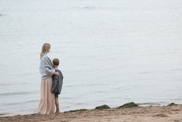 A blond mom in a long pink dress is wrapped up in a warm blanket hugging her young son, looking at the sea. view from the back. - Φωτογραφία, εικόνα