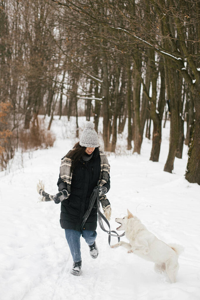 Stylish hipster woman running with cute dog in snowy winter park. Hiking and traveling with pet. Young happy female in cozy clothes having fun with white swiss shepherd dog. Loyal companion - Фото, изображение