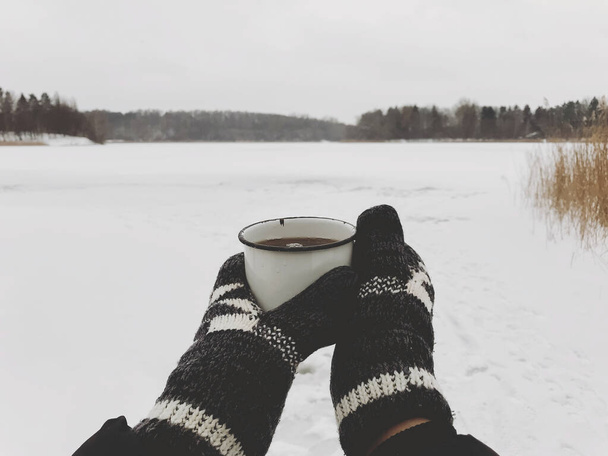 Hands in cozy gloves holding warm cup of tea on background of  snow lake in winter. Hiking and traveling in cold winter season. Warm drink. Space for text. Wanderlust - Photo, Image