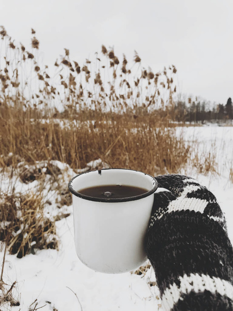 Mano en guante acogedor celebración de taza de té caliente sobre el fondo de la hierba seca y el lago de nieve en invierno. Senderismo y viajes en temporada fría de invierno. Un trago caliente. Espacio para texto - Foto, imagen
