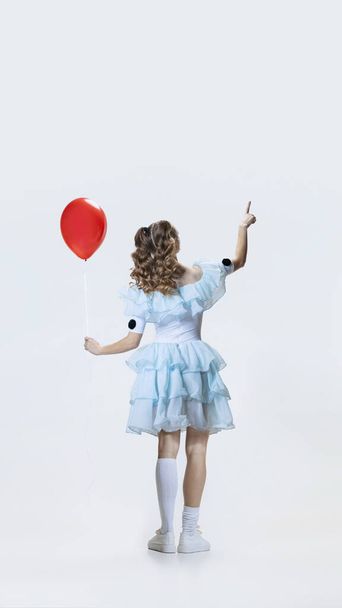 Back view of young girl wearing Halloween dress, costume of movie character standing with red balloon in hand isolated over blue background - Foto, Bild