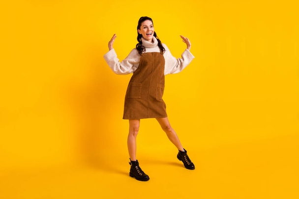 Full length photo of cute adorable young lady dressed brown overall dancing arms hands sides isolated yellow color background - 写真・画像