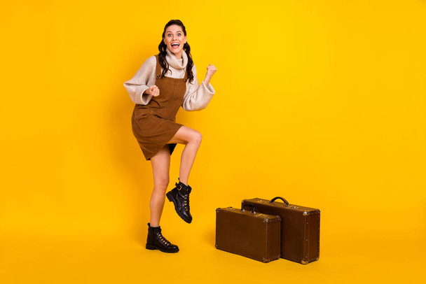 Full length photo of funky lucky young woman wear brown dress baggage rising fists isolated yellow color background - Foto, immagini