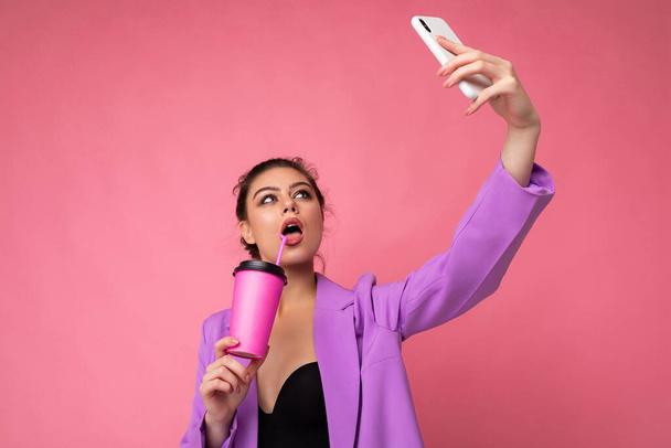 Retrato de cerca de una hermosa mujer morena joven en traje púrpura de moda sobre un fondo rosa en el estudio en una celebración de un teléfono popular y tomar foto selfie y beber a través de una bebida de paja - Foto, Imagen