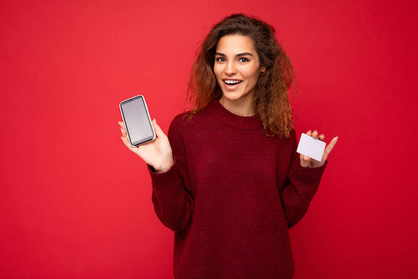 Young beautiful curly brunette woman with sincere emotions poising isolated over background wall with empty space wearing casual dark red sweater holding mobile phone with empty screen display for - Photo, Image