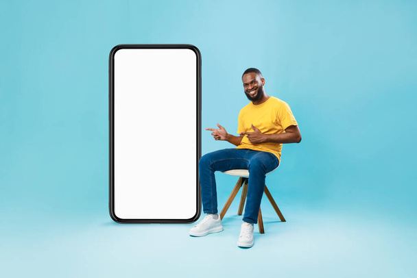 Chico negro feliz sentado en la silla, apuntando a un teléfono inteligente gigante con pantalla blanca, que ofrece maqueta para el diseño de aplicaciones móviles - Foto, imagen