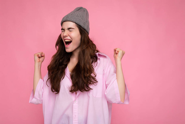 Gyönyörű boldog pozitív fiatal göndör barna nő visel rózsaszín ing és szürke kalap elszigetelt rózsaszín háttér másolás hely kiabálva igen - Fotó, kép