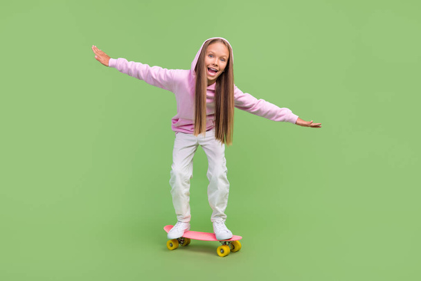 Full body photo of impressed little blond girl ride skate wear hoodie trousers sneakers isolated on green background - Zdjęcie, obraz