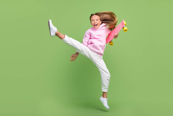 Full body photo of cool small blond girl jump with skate wear hoodie trousers sneakers isolated on green background - Φωτογραφία, εικόνα