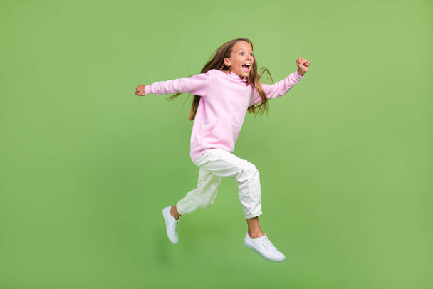 Full length photo of impressed small blond girl run wear sweatshirt trousers sneakers isolated on green background - Zdjęcie, obraz