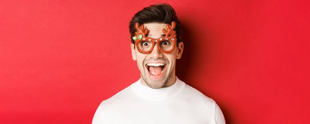 Begrip wintervakantie, kerstmis en viering. Close-up van aantrekkelijke man met haar, het dragen van nieuwjaarsfeest bril en kijken verbaasd over promo aanbod, staande over rode achtergrond - Foto, afbeelding