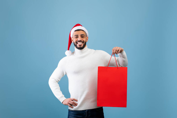 Happy arab man wearing Santa hat and holding shopping bag with Christmas gift over blue background, studio shot - Fotoğraf, Görsel