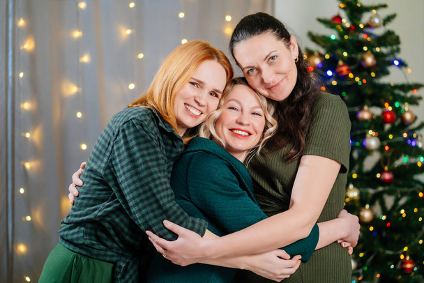 τρεις χαρούμενους και χαρούμενους φίλους αγκαλιάζονται στο χριστουγεννιάτικο δέντρο. γυναικεία φιλία.  - Φωτογραφία, εικόνα