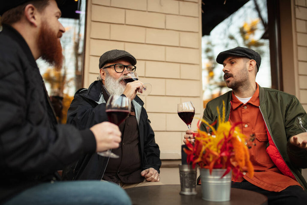 暖かい秋の日にカフェで会う男性の友人のグループ。赤ワインとクリンクグラス - 写真・画像
