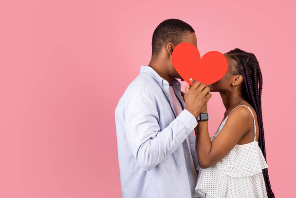 joven romántico negro pareja besos escondiéndose detrás rojo papel corazón - Foto, Imagen