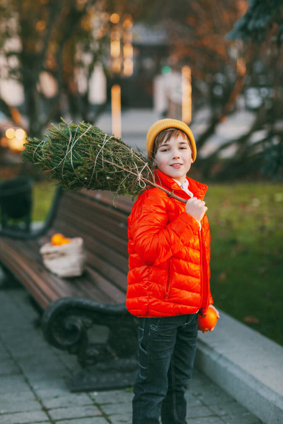 Uroczy chłopiec w pomarańczowej kurtce i kapeluszu spaceruje po parku z choinką na ramieniu. Przygotowania do wakacji, tradycje. Koncepcja Bożego Narodzenia i Nowego Roku - Zdjęcie, obraz