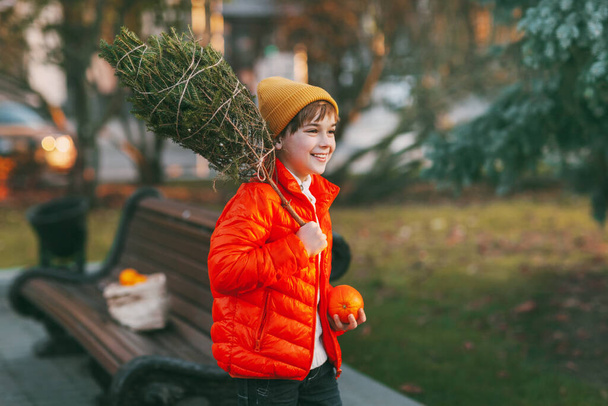 Omzunda Noel ağacı olan turuncu ceketli ve şapkalı sevimli bir çocuk parkta yürüyor. Tatil için hazırlık, gelenekler. Noel ve Yeni Yıl kavramı - Fotoğraf, Görsel