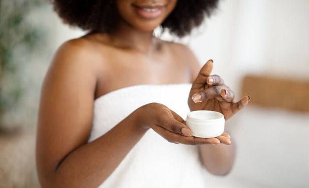 Vista cortada da jovem afro-americana usando creme hidratante, usando toalha de banho dentro de casa, close-up - Foto, Imagem