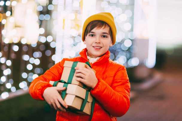 Счастливый мальчик в оранжевой куртке с подарочной коробкой в руках на рождественском базаре зимним вечером. Рождественские подарки, подготовка к праздникам - Фото, изображение