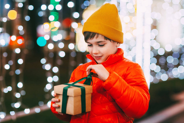 Portrét šťastného chlapce stojícího na večerní ulici a otevírajícího vánoční dárek. Vánoční dárky, sváteční atmosféra. Koncept Vánoc a Nového roku - Fotografie, Obrázek