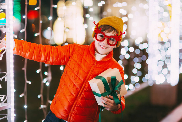 Веселый мальчик в оранжевой куртке и смешных очках ходит по вечернему городу с праздничным освещением. Рождественские подарки, подготовка к праздникам - Фото, изображение