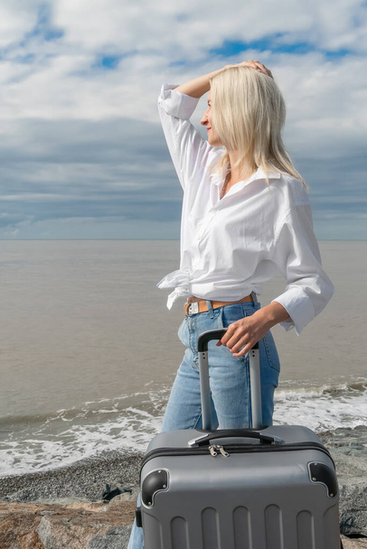 Nuori blondi nainen, jolla on kohotettu käsi päänsä päällä ja hymy, farkut ja paita, jossa on harmaa matkalaukku, seisoo meren rannalla ja katsoo etäisyyteen pilvisellä säällä. Pystykuva - Valokuva, kuva