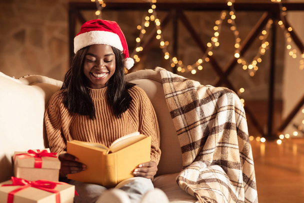 Attractive millennial black lady in Santa hat reading book on Christmas eve in decorated living room, copy space - Foto, afbeelding