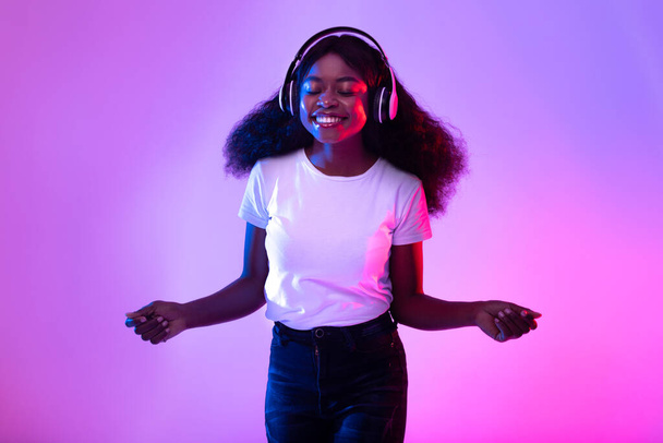 Retrato de alegre jovem negra usando fones de ouvido sem fio, ouvindo música em luz de néon - Foto, Imagem