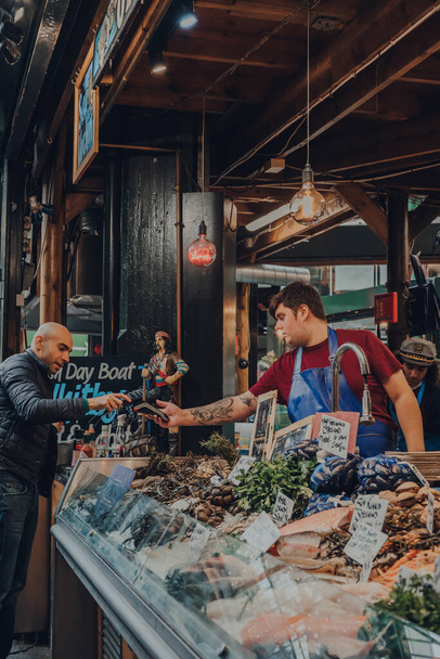 Londyn, Wielka Brytania - 17 października 2021: Klient płacący za zakup w stoisku sprzedawców ryb Shellseekers w Borough Market, jednym z największych i najstarszych rynków żywności w Londynie, z telefonem komórkowym. - Zdjęcie, obraz