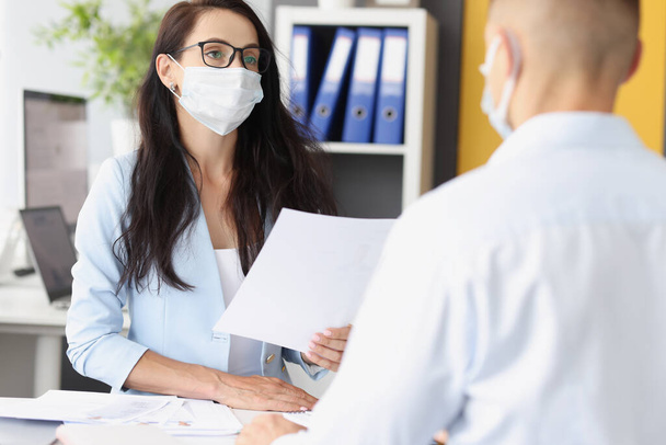 Üzletasszony védő orvosi maszkban dokumentumokat tart és beszél az irodában lévő férfival. - Fotó, kép