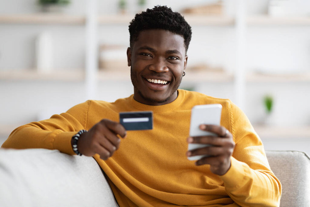 Ευτυχισμένος νεαρός μαύρος που χρησιμοποιεί κινητό και κρατάει πιστωτική κάρτα. - Φωτογραφία, εικόνα