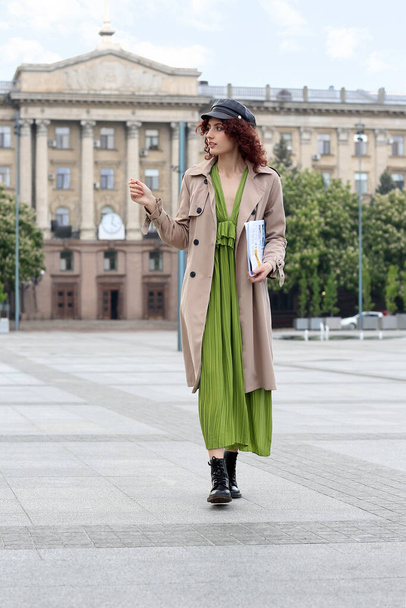 Jovem mulher em boné de couro segurando jornal e andando na praça da cidade - Foto, Imagem