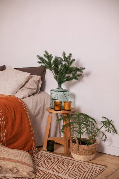 Modern room decor for christmas with bed and vase with branches pine on wooden table. Hygge Xmas decoration. Cozy rustic home. Scandinavian bedroom interior room. Basket with fir tree at living room. - Photo, Image