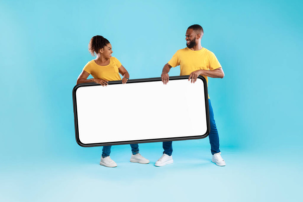 Θυμωμένο νεαρό μαύρο ζευγάρι που μαλώνει για ένα μεγάλο smartphone με λευκή οθόνη σε μπλε φόντο στούντιο, mockup - Φωτογραφία, εικόνα