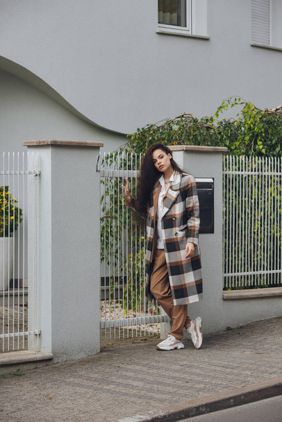 Closeup portrait of young beautiful fashionable woman wearing checkered long coat, beige pants and white blouse . Lady posing on city street. - Фото, изображение